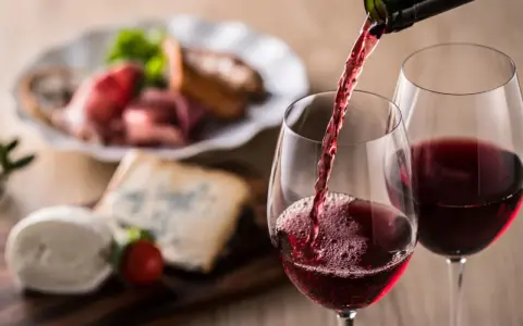 Rött vin hälls upp i glas, perfekt vin till grillat. Ost och charkuterier i bakgrunden.