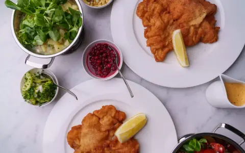 Två krispiga schnitzlar med citronklyfta, serverade med potatissallad, lingon, gurksallad och en krämig sås. Ett perfekt schnitzel recept för en festmåltid.