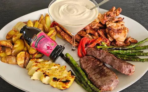 En festlig tallrik med grillad biff, kyckling, halloumi, potatis, sparris och en krämig sås. En flaska "Touch of Taste" sås till grillat står lutad mot potatisen.