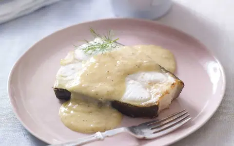 En bit fisk täckt med krämig beurre blanc-sås, garnerad med dill, serveras på en ljusrosa tallrik med en gaffel.