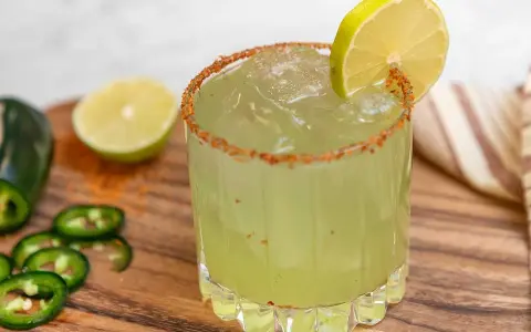 En glas med en kryddig margarita, garnerad med lime och chilipulver. Bredvid ligger jalapeños och limeklyftor.