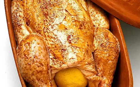En hel kyckling med gyllene skinn och en citron i magen, redo att tillagas i en lergryta.