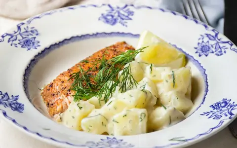 En bit lax med dill och en klyfta citron serveras med krämig stuvad potatis på en vacker blåvit tallrik.