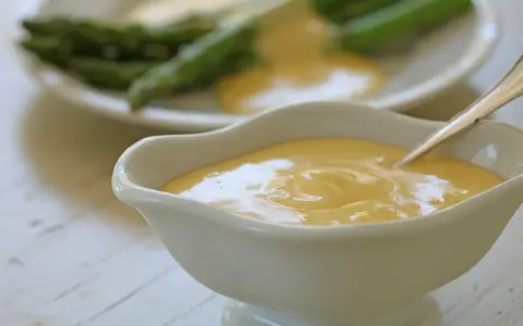 Krämig hollandaisesås i ett serveringskärl, perfekt för ditt hollandaisesås recept. Sparris syns i bakgrunden.
