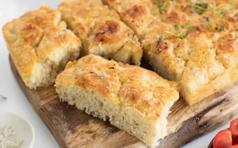 En gyllene focaccia långpanna, skuren i bitar, toppad med örter och flingsalt, ligger på en träskärbräda.