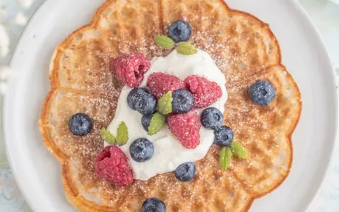 En vacker, blomformad våffla toppad med vispgrädde, hallon och blåbär. Perfekt för en fika med glutenfria våfflor.
