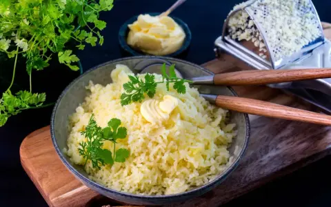 En skål med fluffig, pressad potatis toppad med smör och färska örter. Bredvid syns en smörskål och en rivjärn.