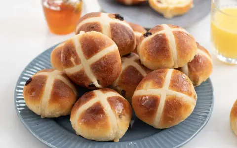 En hög med gyllene, nybakade hot cross buns på en blå tallrik, redo att avnjutas med honung och apelsinjuice.