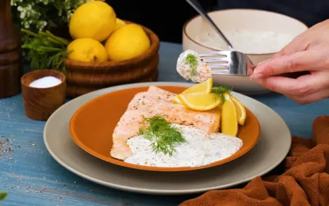 En bit lax med citronskivor och dill, toppad med en krämig romsås till lax. En gaffel med romsås lyfts från skålen.