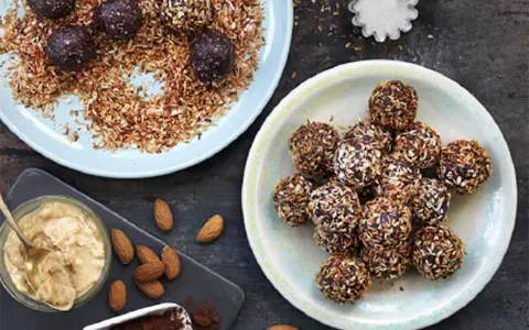 Dadelbollar recept: Mörka, kokosöverdragna energibollar på en ljusblå tallrik, bredvid en skål med mandelsmör och kakaopulver.