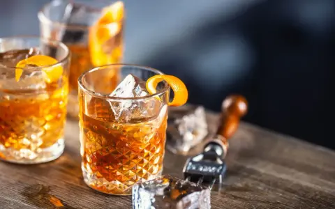 Tre old fashioned-drinkar med is och apelsinskal på ett träbord. En cocktailgaffel syns i bakgrunden.