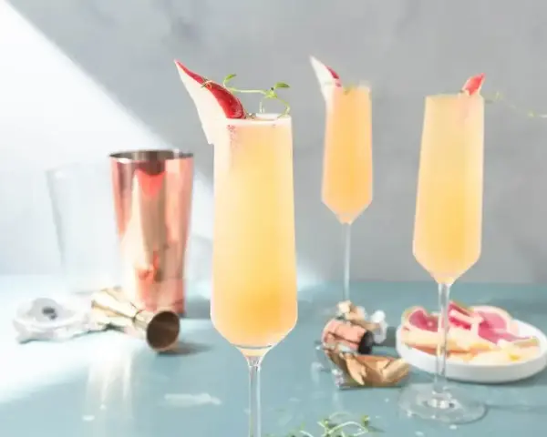 Tre eleganta glas med en ljusgul drink med gin och prosecco, garnerade med en tunn skiva röd frukt och en kvist.