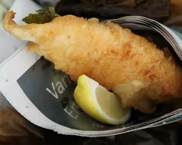 Krispigt friterad fisk med citronklyfta, inslagen i tidningspapper. En klassisk fish and chips-upplevelse.