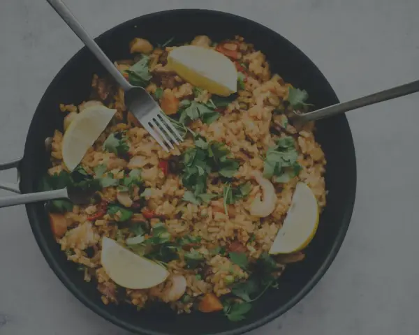 En stor panna med jollof rice, räkor, citronklyftor och färsk persilja. Tre bestick ligger redo att avnjutas.