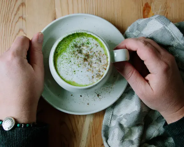 Händerna håller en kopp med en krämig matcha latte, toppad med skum och lite kryddor. En mysig stund med en grön dryck.
