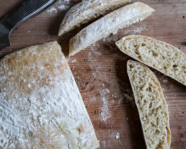 En hel och skivad ciabatta på en träskärbräda, redo för ett gott ciabatta recept. Mjöl strött över.