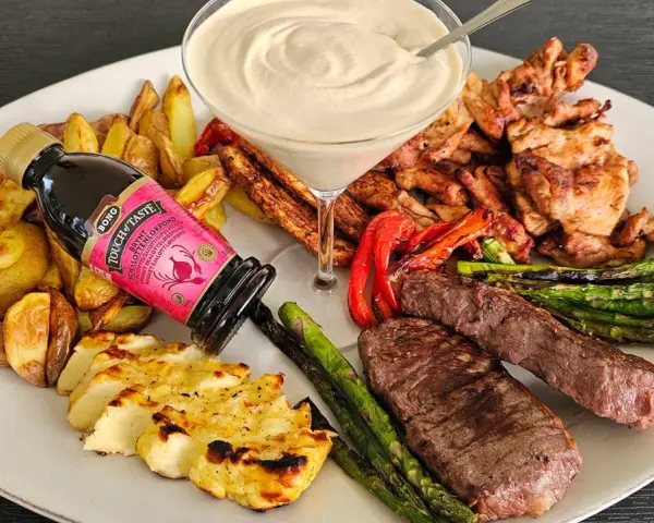 En festlig tallrik med grillad biff, kyckling, halloumi, potatis, sparris och en krämig sås. En flaska "Touch of Taste" sås till grillat står lutad mot potatisen.