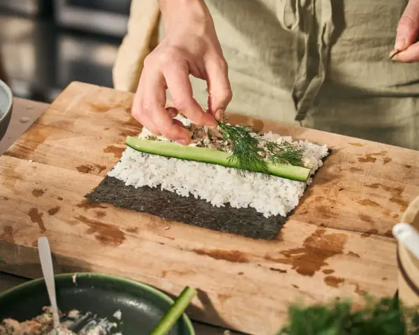 Händer lägger dill på hemmagjord sushi med ris, gurka och fyllning på en skärbräda.
