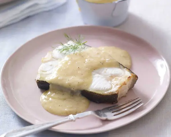En bit fisk täckt med krämig beurre blanc-sås, garnerad med dill, serveras på en ljusrosa tallrik med en gaffel.
