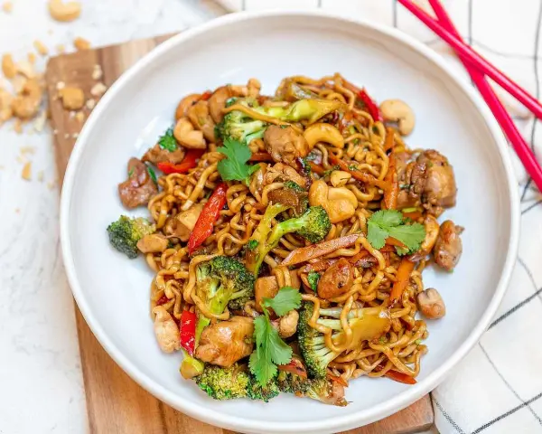 En skål med nudlar, kyckling, broccoli, paprika och cashewnötter. En perfekt allt i ett mat-rätt, redo att ätas med röda ätpinnar.