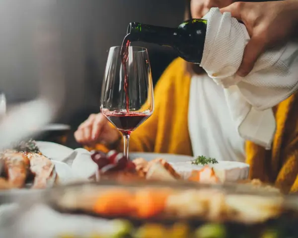 Rödvinet hälls upp i ett glas vid en middagsbjudning. En person i gul tröja sitter bakom. Frågan om hur mycket vin per person är relevant för en trevlig kväll.