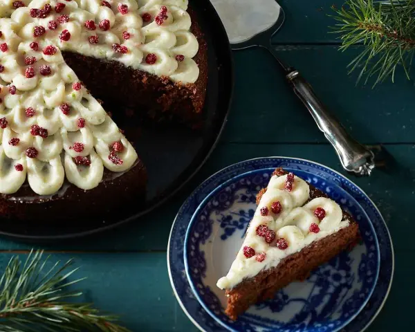 En mjuk pepparkaka med fluffig frosting och röda bär. En bit är upplagd på en blåvit tallrik, redo att avnjutas.
