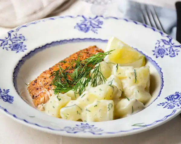 En bit lax med dill och en klyfta citron serveras med krämig stuvad potatis på en vacker blåvit tallrik.