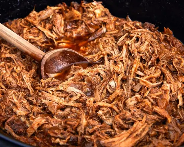 En stor gryta med saftig, långkokt pulled pork i en mörk sås. En träsked vilar i den läckra pulled pork grytan.