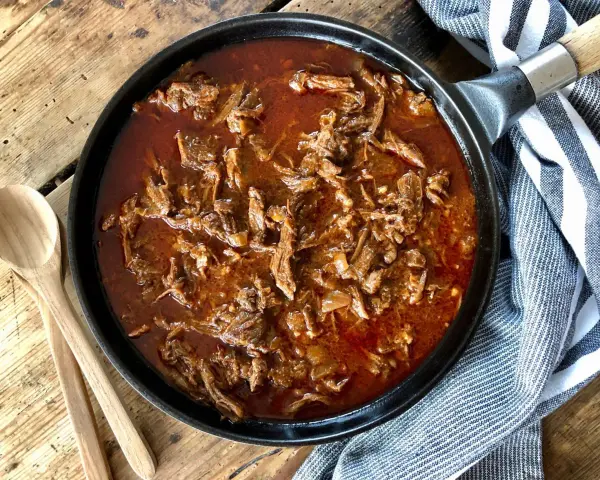 En gjutjärnspanna fylld med saftig, långkokt pulled beef i en mustig sås. Två träskedar ligger bredvid, redo att servera.