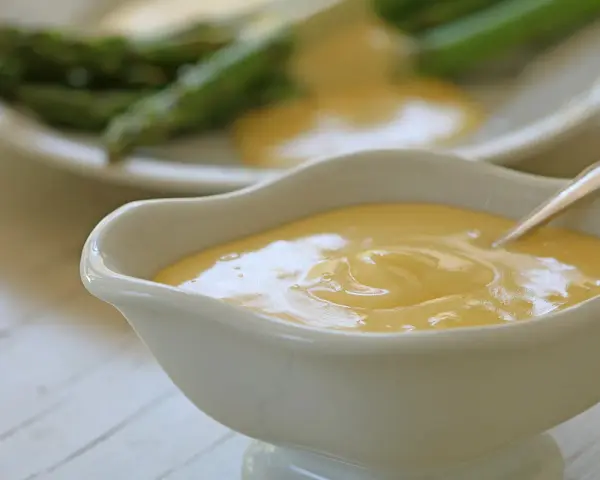 Krämig hollandaisesås i ett serveringskärl, perfekt för ditt hollandaisesås recept. Sparris syns i bakgrunden.