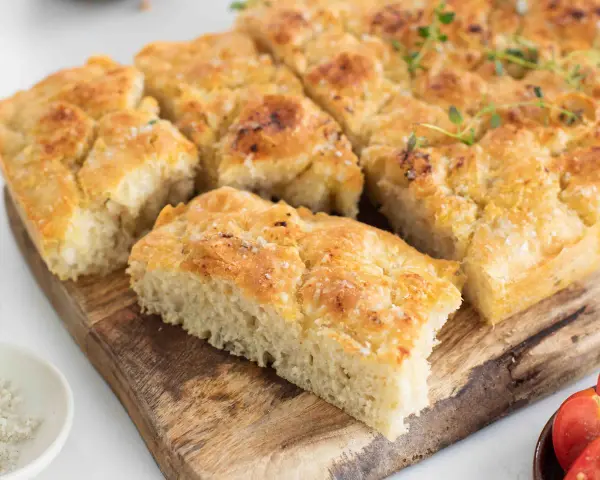 En gyllene focaccia långpanna, skuren i bitar, toppad med örter och flingsalt, ligger på en träskärbräda.