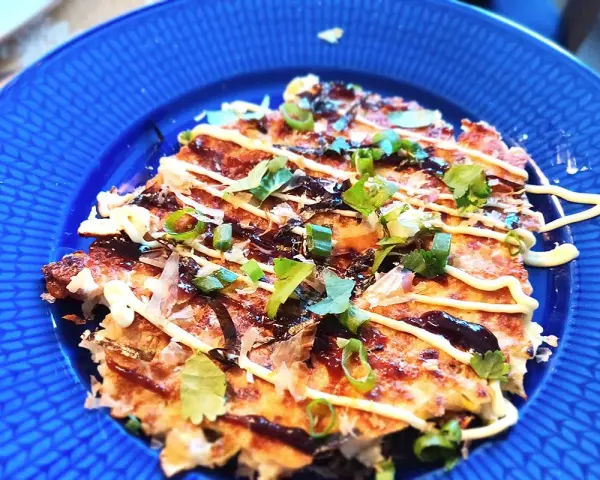 En läcker okonomiyaki på en blå tallrik, toppad med majonnäs, salladslök och torkad sjögräs. Ett perfekt okonomiyaki recept!