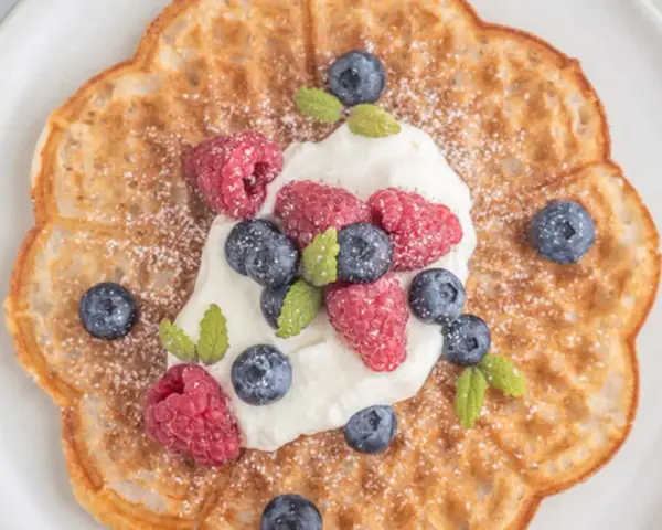 En vacker, blomformad våffla toppad med vispgrädde, hallon och blåbär. Perfekt för en fika med glutenfria våfflor.