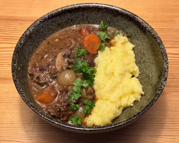 En skål med mustig biff bourguignon med möra köttbitar, champinjoner, lök och morötter, serverad med krämigt potatismos och garnerad med persilja.