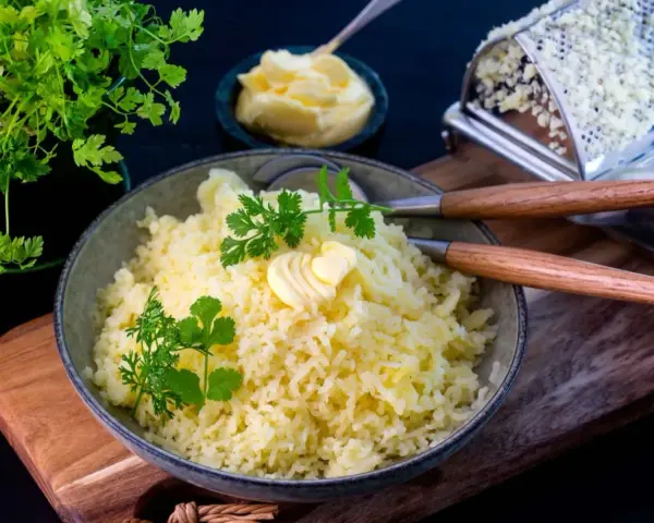 En skål med fluffig, pressad potatis toppad med smör och färska örter. Bredvid syns en smörskål och en rivjärn.