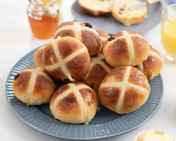 En hög med gyllene, nybakade hot cross buns på en blå tallrik, redo att avnjutas med honung och apelsinjuice.