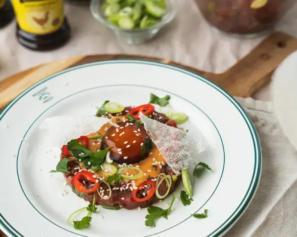 En läcker asiatisk råbiff med chili, salladslök och sesamfrön, serverad på en vit tallrik med gröna kanter.