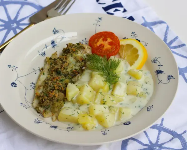 En tallrik med panerad fisk, dillstuvad potatis, en tomatklyfta och en citronklyfta. Allt serveras på en tallrik med blått mönster.