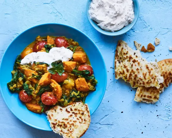 En blå skål med en mustig daal med sötpotatis, spenat och tomater, toppad med gräddfil. Serveras med naanbröd.