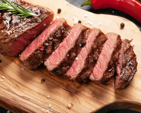 Perfekt stekt entrecote, skivad och serverad med rosmarin, pepparkorn och flingsalt på en träskärbräda. En röd chili ligger bredvid.
