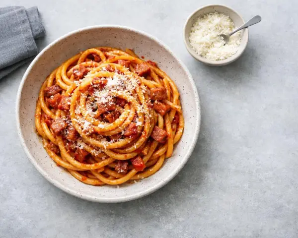 En skål med rörig pasta amatriciana, toppad med riven ost. En liten skål med mer ost och en grå servett syns bredvid.