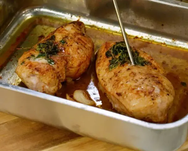 Två saftiga, gyllene grillade kycklingbröst i en ugnsform med örter och vitlök. En termometer mäter temperaturen på den ena kycklingen.