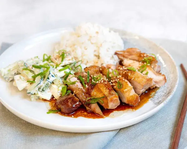 En tallrik med saftig chicken teriyaki, ris och en krämig gurksallad. Garnerad med salladslök och sesamfrön.