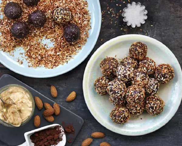Dadelbollar recept: Mörka, kokosöverdragna energibollar på en ljusblå tallrik, bredvid en skål med mandelsmör och kakaopulver.