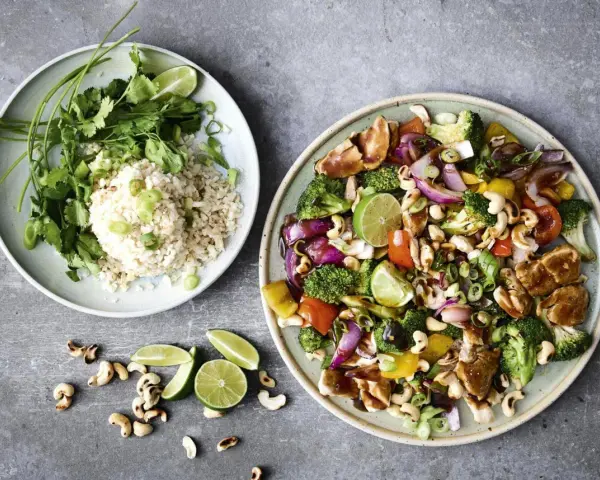 Två tallrikar med mat. En med ris, koriander och salladslök. Den andra med kyckling, broccoli, paprika, cashewnötter och lime. Perfekt för nyttiga recept.