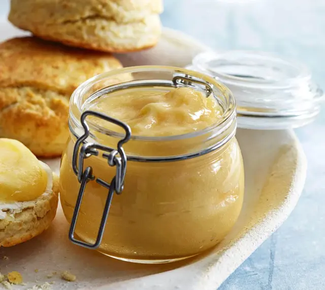 En burk med gyllene lemon curd bredvid scones, en smörgås med grädde och lemon curd.