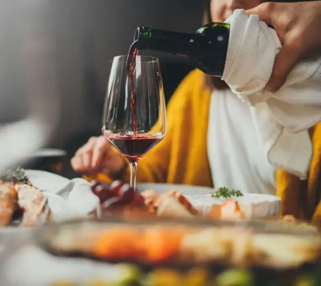 Rödvinet hälls upp i ett glas vid en middagsbjudning. En person i gul tröja sitter bakom. Frågan om hur mycket vin per person är relevant för en trevlig kväll.