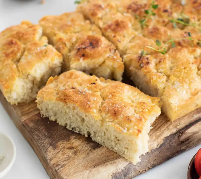 En gyllene focaccia långpanna, skuren i bitar, toppad med örter och flingsalt, ligger på en träskärbräda.