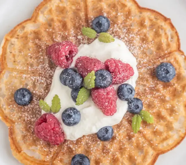En vacker, blomformad våffla toppad med vispgrädde, hallon och blåbär. Perfekt för en fika med glutenfria våfflor.