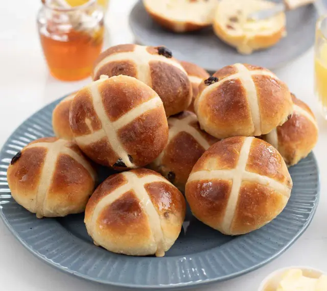 En hög med gyllene, nybakade hot cross buns på en blå tallrik, redo att avnjutas med honung och apelsinjuice.